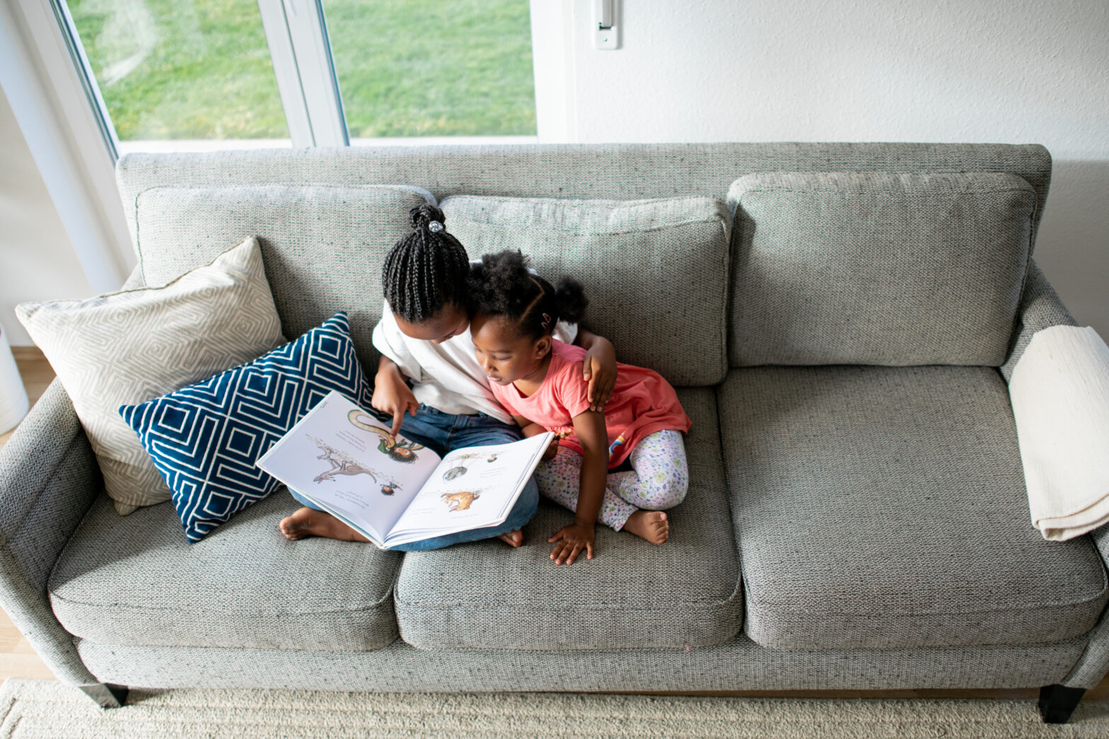 Two sisters sit cross-legged on a grey couch reading a book