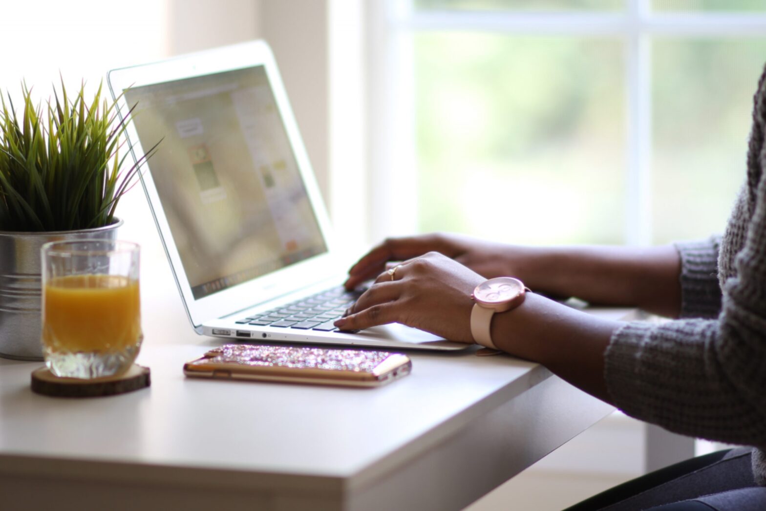 A Black woman's hands type on a silver laptop in front of a window with trees outside in the background and a glass of orange juice and a green plant and a cell phone on the desk beside the laptop. The woman is wearing a watch and a grey sweater.