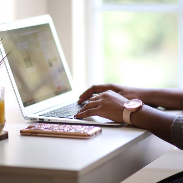 A Black woman's hands type on a silver laptop in front of a window with trees outside in the background and a glass of orange juice and a green plant and a cell phone on the desk beside the laptop. The woman is wearing a watch and a grey sweater.