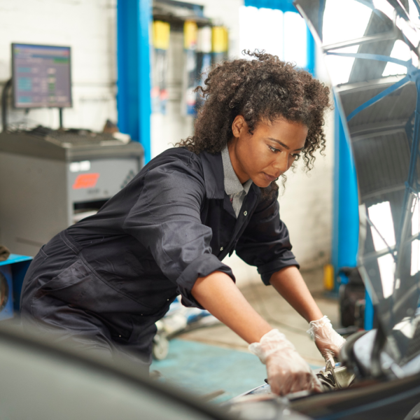 A Black woman wearing navy coveralls and white gloves bends over the open hood of a car to perform mechanic maintenance. Tools and mechanic equipment are visible in the background.