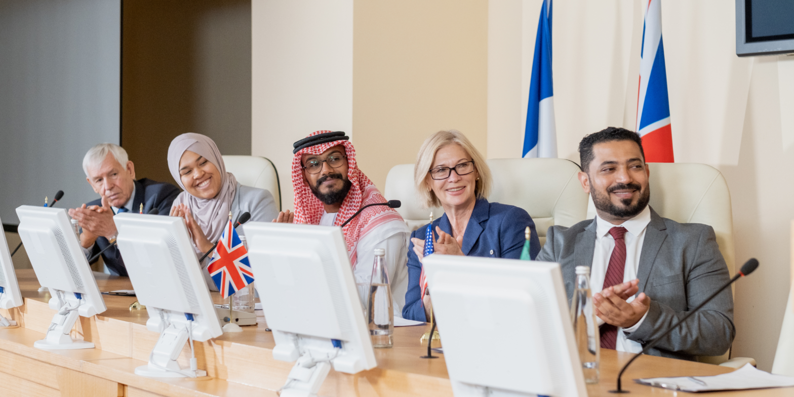 Five people from different cultural backgrounds sit together at a long wooden table in a meeting room. White desktop computers sit in front of each person, with two flags against the wall behind the table. The countries of the flags are not visible.