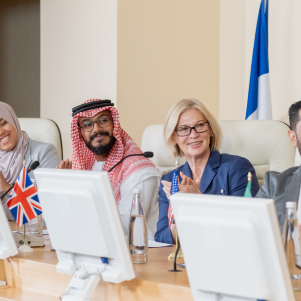 Five people from different cultural backgrounds sit together at a long wooden table in a meeting room. White desktop computers sit in front of each person, with two flags against the wall behind the table. The countries of the flags are not visible.