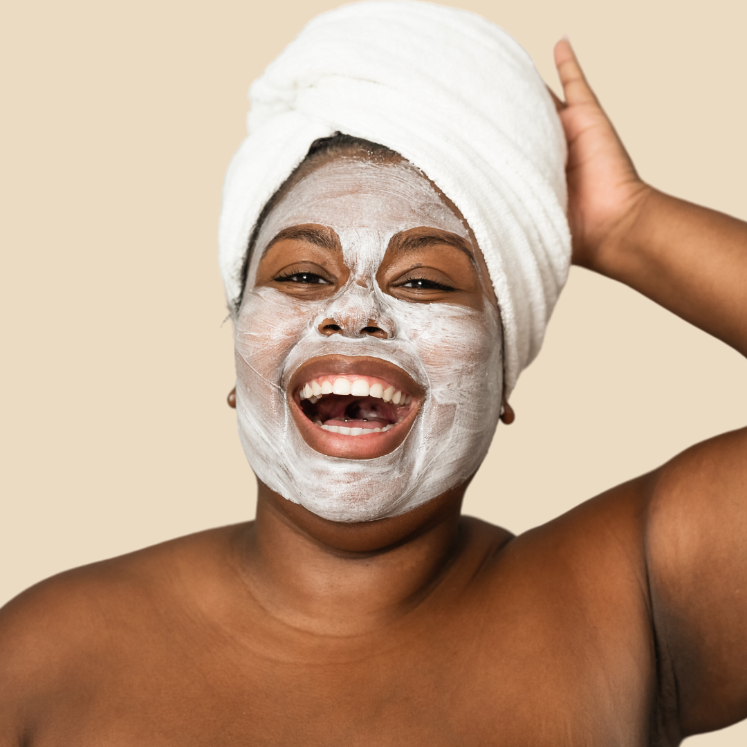 A Black, plus-size woman laughs heartily into the screen while wearing a white clay face mask and a towel in a turban on her head, evoking an image of a day spa.