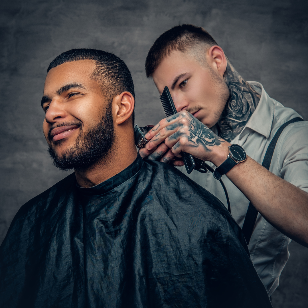 A Latino male barber with tatoo body art visible on hands and neck trims the hair of a Black man sitting in front of him wearing a black barber's cape.