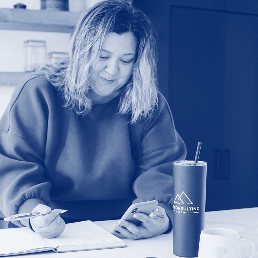A business woman with shoulder-length hair leans over a counter with pen in one hand and phone in the other, about to write notes in a notebook. There is a beverage container with the MT Consulting Group logo in the foreground, as well as a coffee cup.