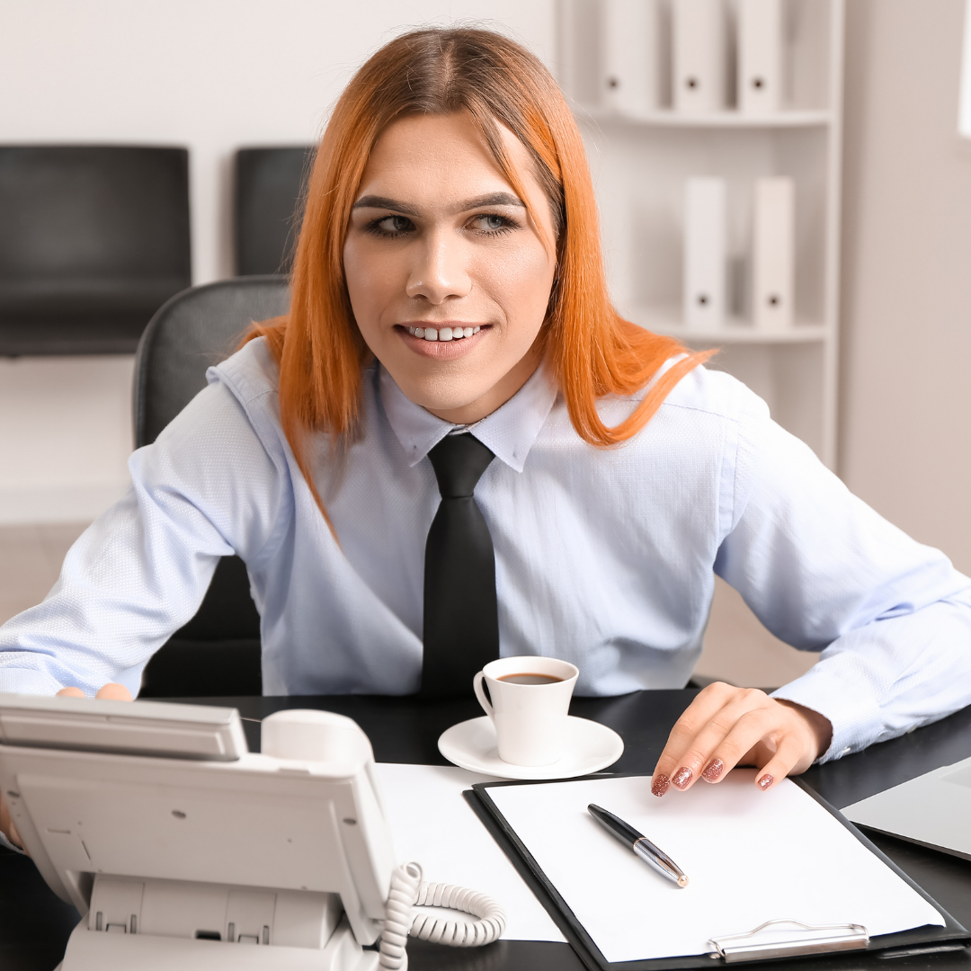 A woman with long red hair and a button up shirt and tie sits at a desk with a pen, notebook and coffee in front of her. With the blog title: How to support gender transitioning employees at work