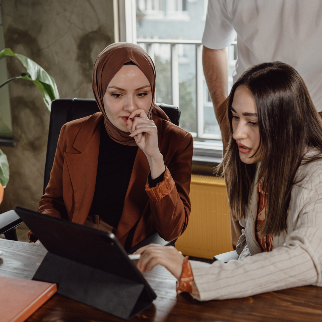 A Chief Diversity Officer, a woman wearing a brown blazer with matching hijab while talking to a woman with long hair. An ipad sits on the desk in front of them wile they review key documents.